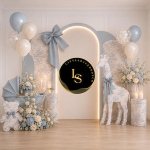 May include: A baby shower backdrop featuring a light blue stroller, teddy bear, and giraffe, all adorned with floral arrangements and bows. Balloons in white and light blue float in the background, with the text "LITTLE BACKDROP CO" on a black circle.
