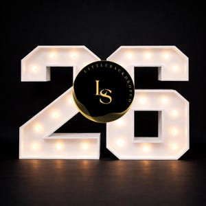 May include: White illuminated marquee numbers 26 with warm white lights. A black circle with gold accents and the text "LITTLE BACKDROP CO" and "ls" is centered on the numbers. The background is black.