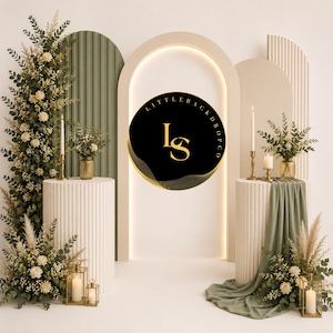 Pode incluir: Um cenário decorativo para eventos com três painéis arqueados em verde sálvia e creme, dois pedestais cilíndricos canelados e arranjos florais de eucalipto e rosas brancas. Um letreiro circular preto exibe o texto dourado LITTLEBACKDROPCO LS.