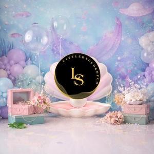 May include: A whimsical backdrop featuring a mermaid theme with a large seashell, balloons, and pastel colors. Pink and blue treasure chests are stacked with floral arrangements. The background has a dreamy underwater scene with fish and bubbles.