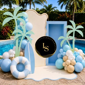 May include: A beach-themed party backdrop with light blue, white, and beige elements. Features palm tree cutouts, a surfboard, a life preserver, and balloon clusters. The backdrop includes the text "LITTLE BACKDROP CO" in a circular logo.