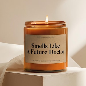 May include: A lit orange candle in a clear glass jar with a brown label that reads "Smells Like A Future Doctor." The candle is labeled "Whisper & Flame Candle" and is hand-poured with 100% soy wax, 9 oz.