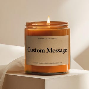 May include: A lit orange candle in a clear glass jar with a brown label that reads "Custom Message." The candle is labeled "Whisper & Flame Candle" and is 9 oz. of 100% soy wax.