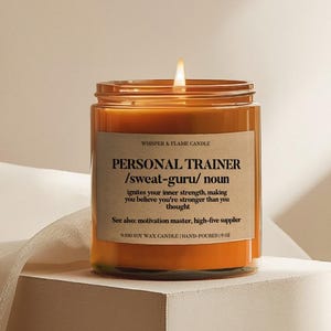 May include: An orange-coloured candle in a clear glass jar with a lit flame. The label says "PERSONAL TRAINER /sweat-guru/ noun". The candle is made from soy wax and is 9 oz. The background is a neutral colour.