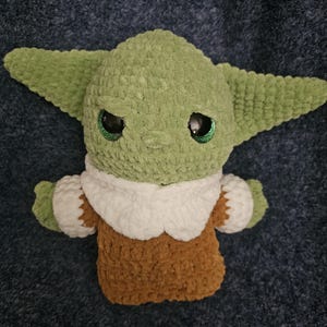 May include: A handmade green crocheted plush toy with large ears and large black eyes. The toy is wearing a brown and white outfit. The plush toy is a character from a popular science fiction franchise.
