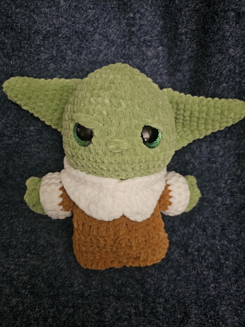 May include: A handmade green crocheted plush toy with large ears and large black eyes. The toy is wearing a brown and white outfit. The plush toy is a character from a popular science fiction franchise.