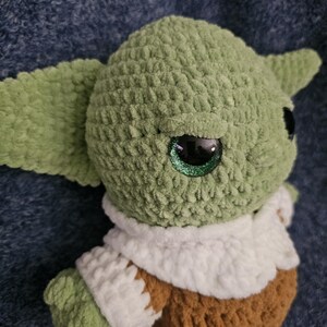 May include: A handmade green crocheted doll with large black eyes and pointed ears. The doll is wearing a white and brown outfit. The doll's eyes have a glittery green sparkle. The doll is a character from a popular science fiction franchise.