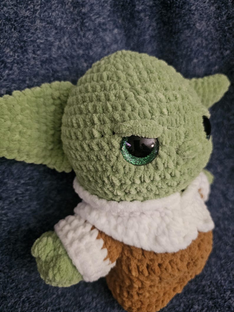 May include: A handmade green crocheted doll with large black eyes and pointed ears. The doll is wearing a white and brown outfit. The doll's eyes have a glittery green sparkle. The doll is a character from a popular science fiction franchise.