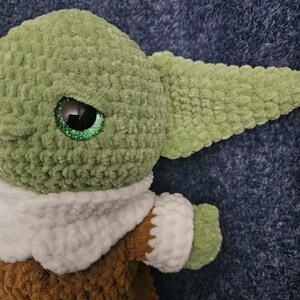 May include: A handmade plush doll of a character with large ears and big eyes. The doll is primarily green, with a brown outfit and white collar. The eyes have a glittery effect. The doll is made of a soft, textured material.