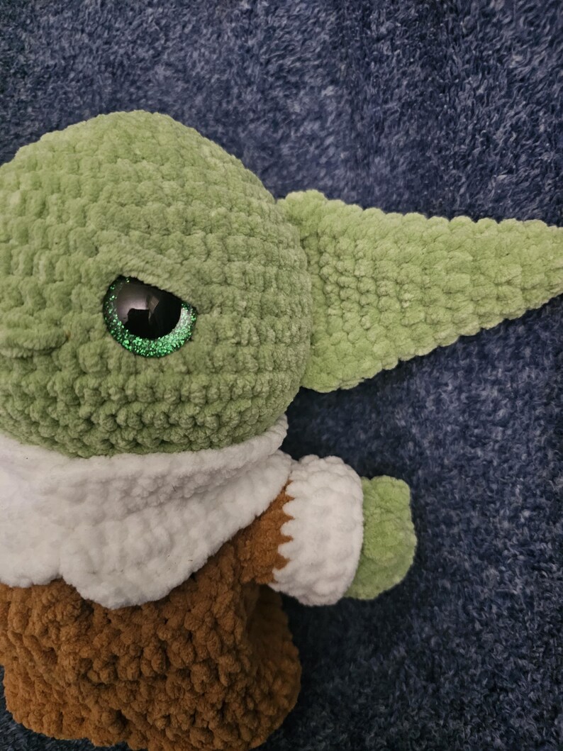 May include: A handmade plush doll of a character with large ears and big eyes. The doll is primarily green, with a brown outfit and white collar. The eyes have a glittery effect. The doll is made of a soft, textured material.