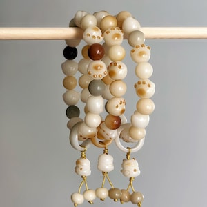 May include: Three beaded bracelets with a variety of off-white, brown, and gray beads. Some beads are paw prints. Each bracelet has a white ring and dangling charms with gold string and small beads. The bracelets are hanging on a wooden dowel.