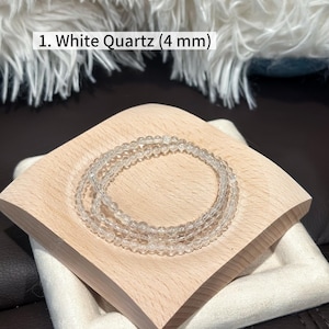 May include: Three clear white quartz bracelets, each with 4 mm beads, are displayed on a light wooden square tray. The tray sits atop a white fabric-covered base, with a soft, white, fluffy background.