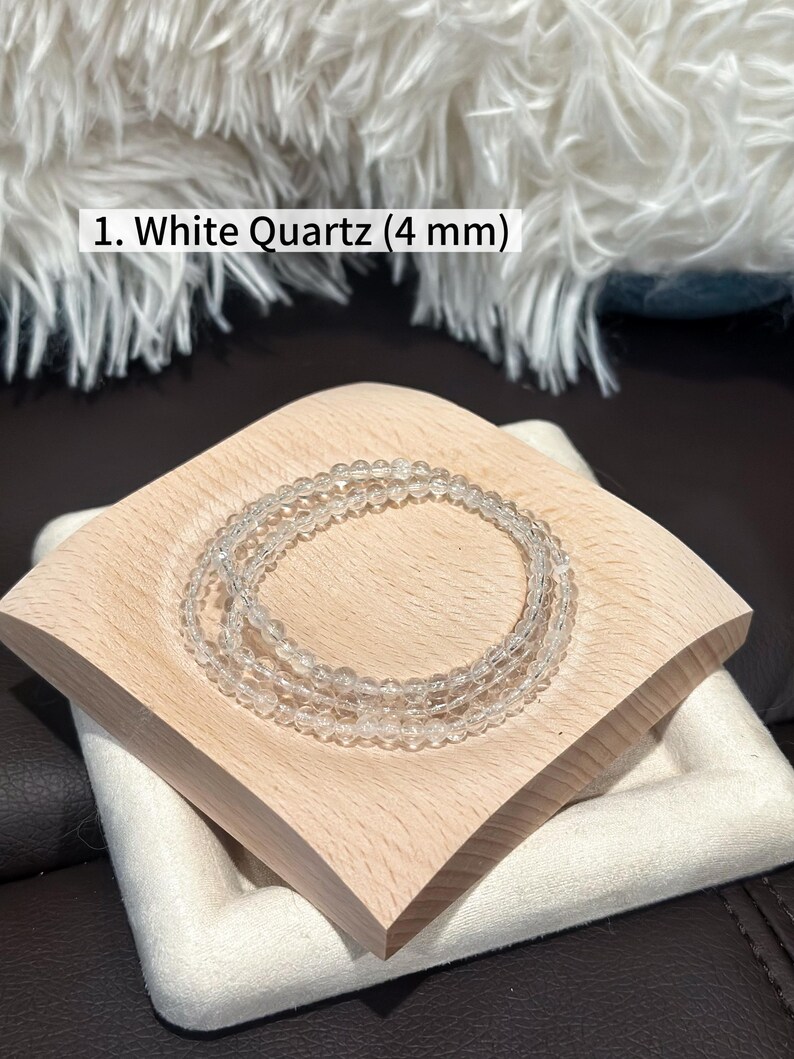 May include: Three clear white quartz bracelets, each with 4 mm beads, are displayed on a light wooden square tray. The tray sits atop a white fabric-covered base, with a soft, white, fluffy background.