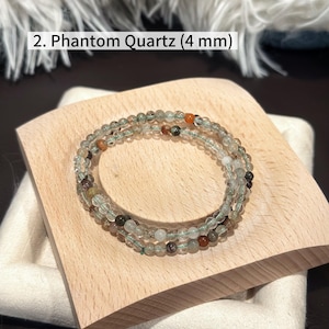 May include: A Phantom Quartz bracelet with 4 mm beads, displayed on a wooden block. The bracelet features clear and green beads with hints of orange and brown. Text in the image reads: "2. Phantom Quartz (4 mm)".