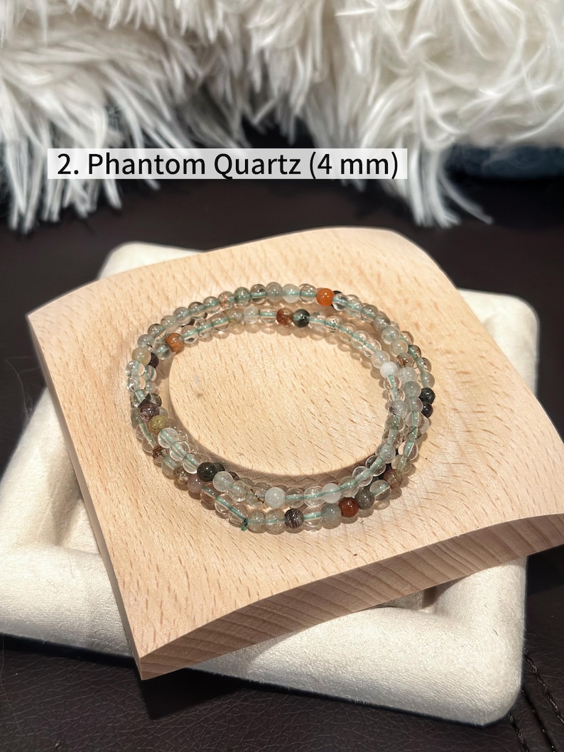 May include: A Phantom Quartz bracelet with 4 mm beads, displayed on a wooden block. The bracelet features clear and green beads with hints of orange and brown. Text in the image reads: "2. Phantom Quartz (4 mm)".