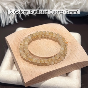 May include: A golden rutilated quartz bracelet with 6 mm beads. The bracelet is displayed on a small, light-colored wooden block. The text in the image reads: 6. Golden Rutilated Quartz (6 mm).