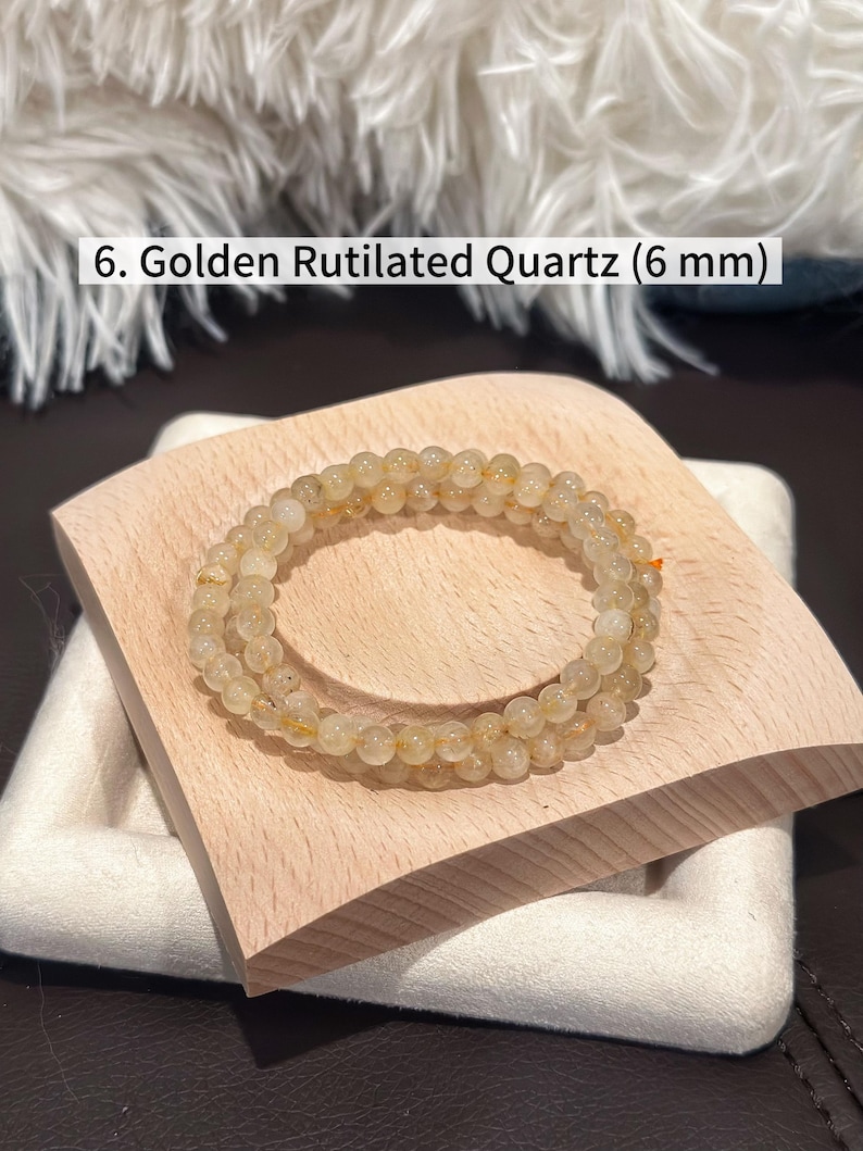 May include: A golden rutilated quartz bracelet with 6 mm beads. The bracelet is displayed on a small, light-colored wooden block. The text in the image reads: 6. Golden Rutilated Quartz (6 mm).