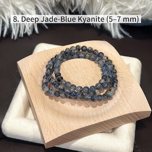 May include: Deep jade-blue kyanite beaded bracelets, each bead measuring 5-7 mm. The bracelets are displayed on a light wood block, showcasing the natural color variations of the stones. The text "8. Deep Jade-Blue Kyanite (5-7 mm)" is visible.