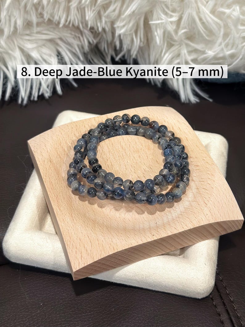 May include: Deep jade-blue kyanite beaded bracelets, each bead measuring 5-7 mm. The bracelets are displayed on a light wood block, showcasing the natural color variations of the stones. The text "8. Deep Jade-Blue Kyanite (5-7 mm)" is visible.