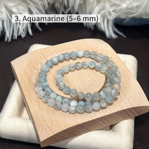 May include: Aquamarine beaded bracelet, featuring 5-6 mm beads. The bracelet is displayed on a light wooden tray with a white fabric base. The text "3. Aquamarine (5-6 mm)" is visible in the image.