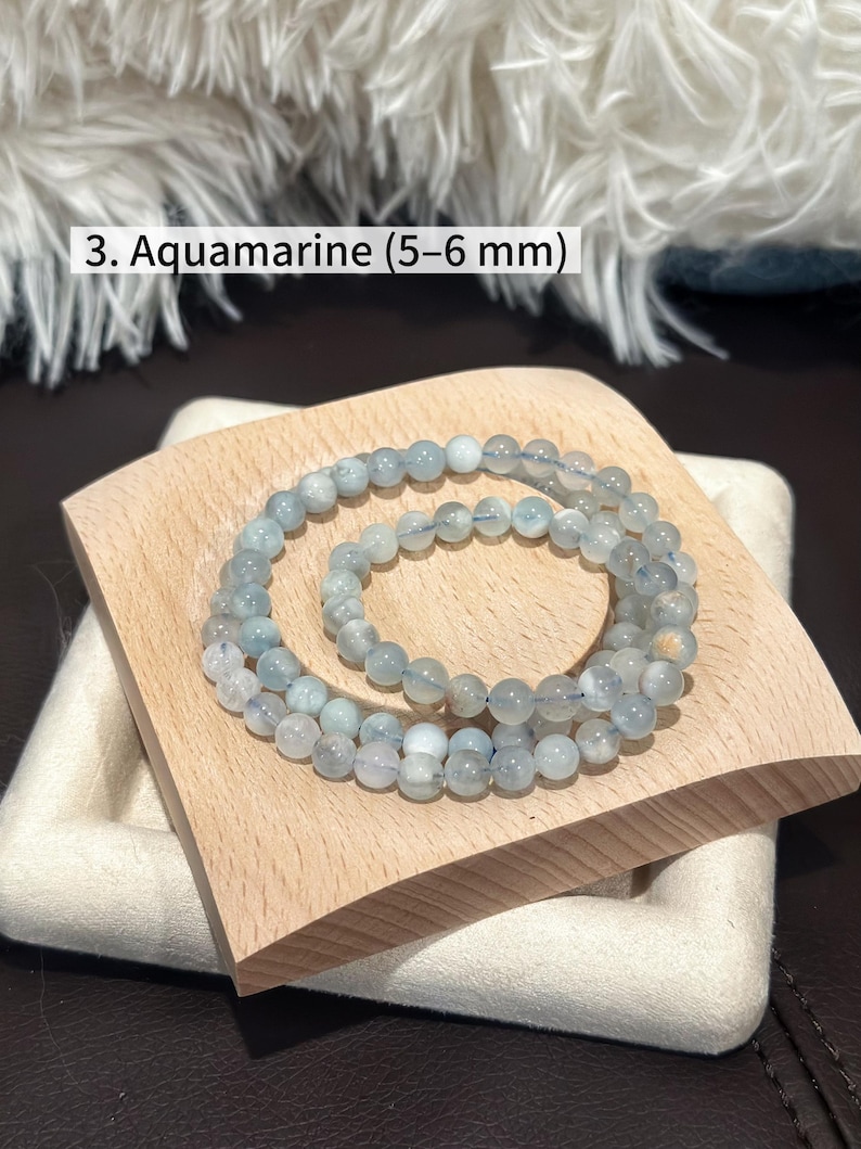 May include: Aquamarine beaded bracelet, featuring 5-6 mm beads. The bracelet is displayed on a light wooden tray with a white fabric base. The text "3. Aquamarine (5-6 mm)" is visible in the image.