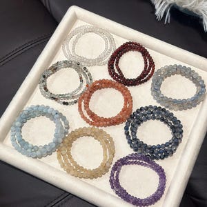 May include: A collection of beaded bracelets in various colors, including clear, red, blue, and purple, arranged in a white tray. Each bracelet appears to be made of small, round beads, and they are stacked in multiple layers.