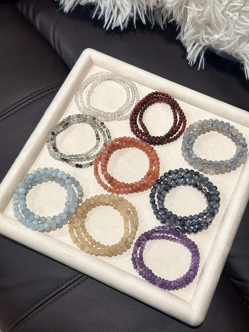 May include: A collection of beaded bracelets in various colors, including clear, red, blue, and purple, arranged in a white tray. Each bracelet appears to be made of small, round beads, and they are stacked in multiple layers.
