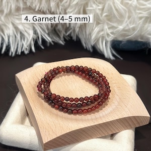 May include: A set of three garnet bead bracelets, each with 4-5 mm beads, displayed on a light wooden stand. The beads are a deep reddish-brown color. The text "4. Garnet (4-5 mm)" is visible in the image.