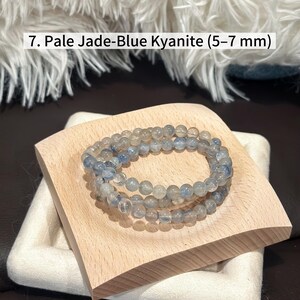 May include: Pale jade-blue kyanite beaded bracelet, featuring 5-7 mm beads. The bracelet is displayed on a light wooden square, resting on a white, textured surface. The background includes a fluffy, white textile.