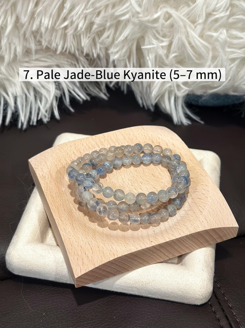 May include: Pale jade-blue kyanite beaded bracelet, featuring 5-7 mm beads. The bracelet is displayed on a light wooden square, resting on a white, textured surface. The background includes a fluffy, white textile.