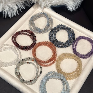 May include: A collection of beaded bracelets in various colors, including clear, burgundy, orange, yellow, blue, and purple. The bracelets are arranged in a tray, showcasing their different hues and textures. The beads appear to be small and round.