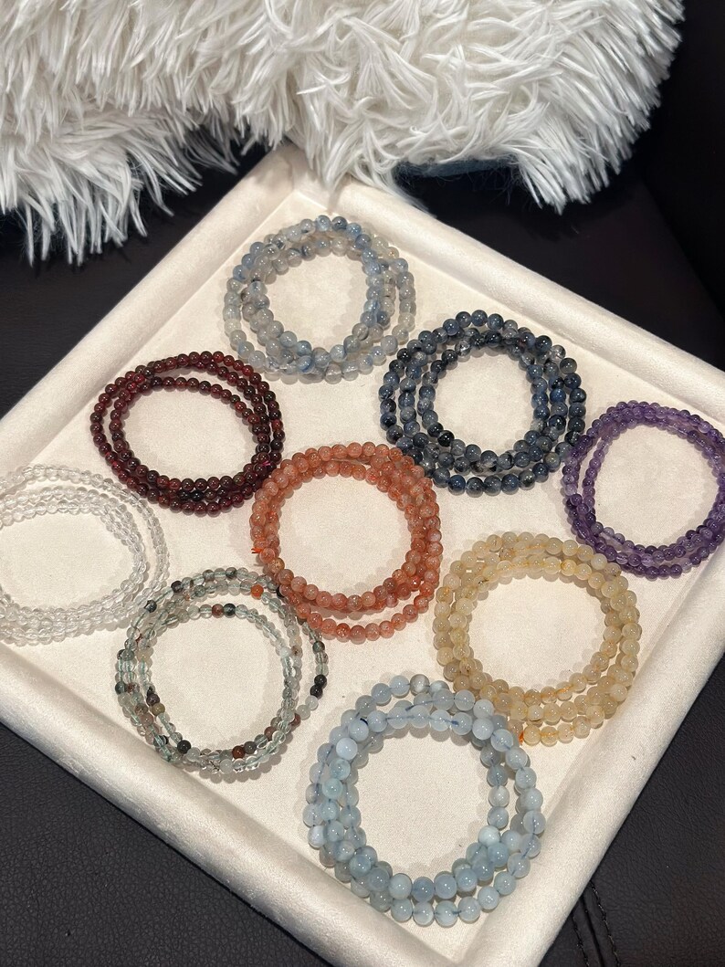 May include: A collection of beaded bracelets in various colors, including clear, burgundy, orange, yellow, blue, and purple. The bracelets are arranged in a tray, showcasing their different hues and textures. The beads appear to be small and round.