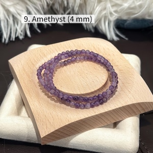 May include: A pair of amethyst bracelets, each composed of small, round, purple beads, are displayed on a light-colored wooden block. The bracelets are approximately 4 mm in diameter. The text "9. Amethyst (4 mm)" is visible in the image.