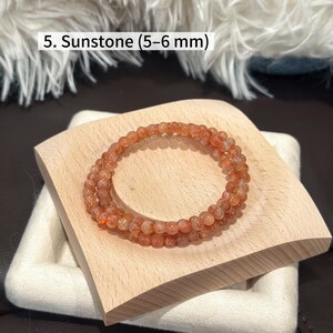 May include: A sunstone bracelet composed of small, round, orange-hued beads, approximately 5-6 mm in diameter. The bracelet is displayed on a light wooden block, with the text "5. Sunstone (5-6 mm)" visible in the image.