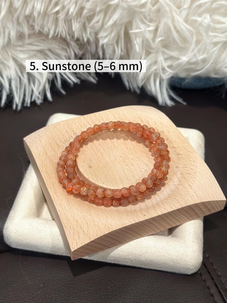 May include: A sunstone bracelet composed of small, round, orange-hued beads, approximately 5-6 mm in diameter. The bracelet is displayed on a light wooden block, with the text "5. Sunstone (5-6 mm)" visible in the image.