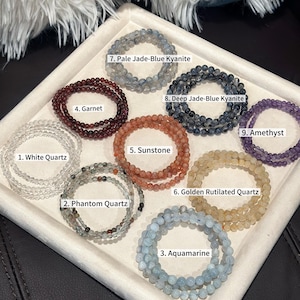 May include: A collection of beaded bracelets in various colors and gemstones, including white quartz, phantom quartz, aquamarine, garnet, sunstone, golden rutilated quartz, pale jade-blue kyanite, deep jade-blue kyanite, and amethyst. Each bracelet is labeled.