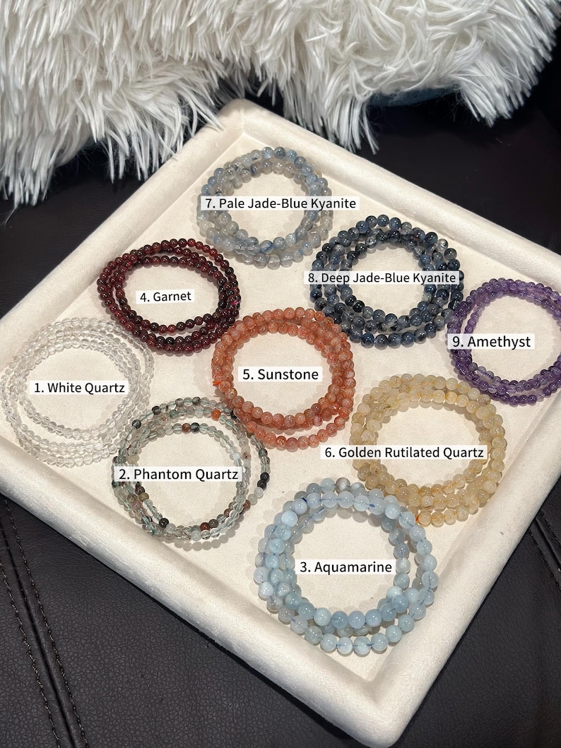 May include: A collection of beaded bracelets in various colors and gemstones, including white quartz, phantom quartz, aquamarine, garnet, sunstone, golden rutilated quartz, pale jade-blue kyanite, deep jade-blue kyanite, and amethyst. Each bracelet is labeled.