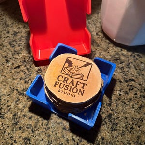 May include: A stack of wooden coasters with the text "CRAFT FUSION STUDIO" in a blue holder, with a red holder in the background. The coasters are round and made of wood. The holders are made of plastic.