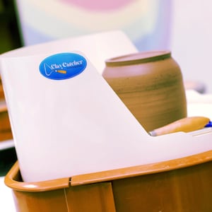 May include: A white plastic clay catcher with a blue oval logo that reads "Clay Catcher". A brown ceramic jar and a wooden tool are visible inside the catcher. The catcher sits in a brown plastic tray.