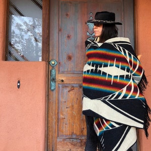 May include: A black hat with a decorative band and a colorful, patterned blanket with fringe. The blanket features a design with white, black, red, and blue stripes. The person is standing in front of a wooden door.