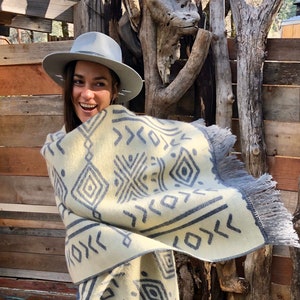 Könnte beinhalten: Eine Frau trägt einen grauen Hut und eine cremefarbene und graue gemusterte Decke mit Fransen. Die Decke hat ein geometrisches Muster. Die Frau steht vor einem Holzzaun.