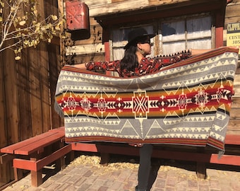 May include: A woman wearing a black hat and a red shirt holds a grey and red patterned blanket with fringe. The blanket has a geometric design with black, red, orange, and yellow stripes.