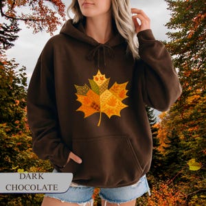 Op de afbeelding: Donkerchocoladebruine hoodie met een groot, kleurrijk esdoornbladontwerp op de voorkant. Het blad bestaat uit verschillende herfstthema's en kleuren. De tekst "DARK CHOCOLATE" staat onderaan.