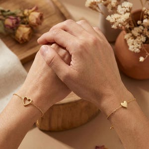 May include: Two gold-colored bracelets, each with a heart-shaped charm, are displayed. One bracelet features a cutout heart, while the other has a solid heart. The bracelets are on wrists, with a neutral background and dried flowers.