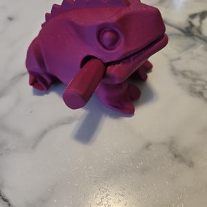 May include: A magenta-colored, 3D-printed lizard toy with a movable jaw and a cylindrical piece extending from its mouth. The lizard has a spiky ridge along its back and is resting on a marble-patterned surface.