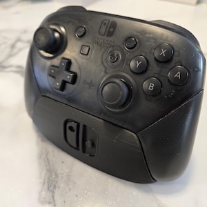 Pode incluir: Controlo Nintendo Switch Pro preto com botões X, Y, A e B. O controlo tem um D-pad, dois joysticks e o logótipo Nintendo Switch. Desenhado para jogos.