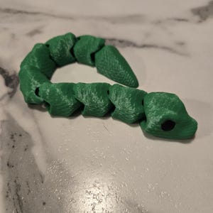 Könnte beinhalten: Ein grünes, bewegliches Schlangenspielzeug. Das Spielzeug besteht aus verbundenen Segmenten, die es ihm ermöglichen, sich zu biegen und zu bewegen. Der Kopf der Schlange hat zwei schwarze Augendetails. Das Spielzeug liegt auf einer weiß-grauen Oberfläche.