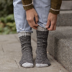 Puede incluir: Un par de calcetines grises y negros a rayas con un puño blanco. Los calcetines están hechos de un material suave y texturizado.