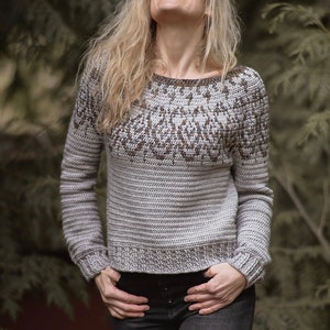 May include: A gray and brown crocheted sweater with a geometric pattern on the neckline. The sweater has long sleeves and a relaxed fit.