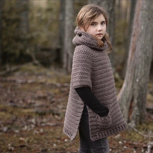 Può includere: Un poncho di uncinetto marrone con collo alto e una tasca sul davanti. Il poncho ha spacco laterale ed è indossato da una persona giovane in un ambiente boschivo.